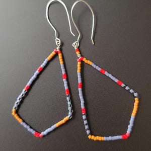 Puede incluir: Un par de pendientes geométricos con cuentas. Los pendientes tienen forma triangular con ganchos plateados. Las cuentas son en tonos grises, naranjas, rojos y negros. Los pendientes se muestran sobre un fondo oscuro.