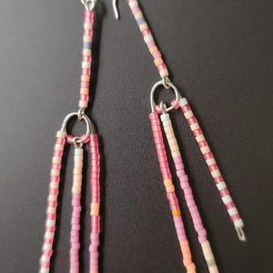 Puede incluir: Pendientes colgantes con ganchos plateados. Los pendientes presentan hebras de cuentas en tonos rosa, blanco y naranja. Las cuentas están dispuestas en un patrón lineal, creando un accesorio delicado y colorido.