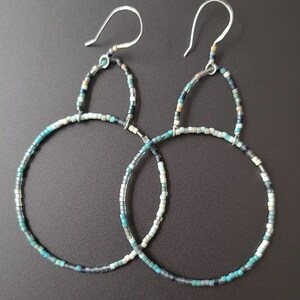 Puede incluir: Un par de pendientes de aro con un diseño de cuentas. Los pendientes tienen un gancho plateado y están adornados con pequeñas cuentas de colores en tonos turquesa, gris y blanco. Los aros son circulares y tienen un estilo minimalista.