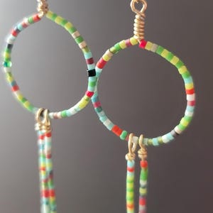 Puede incluir: Un par de pendientes de aro con detalles de cuentas de colores. Los aros están adornados con pequeñas cuentas multicolores en tonos rojos, verdes, amarillos y azules. Se utiliza alambre dorado para conectar los aros y crear hebras de cuentas colgantes.