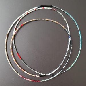 Peut inclure: Trois colliers de perles délicats disposés en couches. Les colliers présentent une variété de couleurs, dont le blanc, le gris, le rouge et le turquoise. Chaque collier est composé de petites perles espacées, créant un design minimaliste et élégant.
