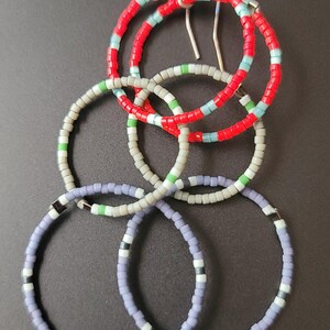 Puede incluir: Pendientes de aro hechos a mano con cuentas en un diseño entrelazado único. Los pendientes presentan cuentas rojas, azul claro, verdes, blancas y lavanda. Los pendientes tienen ganchos plateados y se muestran sobre una superficie gris oscuro.
