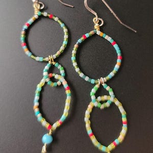 Puede incluir: Pendientes hechos a mano con un diseño colorido. Los pendientes presentan una combinación de formas redondas y ovaladas, conectadas con pequeños eslabones dorados. Cada pendiente tiene una pequeña cuenta azul claro en la parte inferior y ganchos plateados.