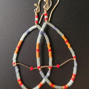 Puede incluir: Pendientes hechos a mano con cuentas con un diseño de lágrima. Los pendientes tienen ganchos dorados y están adornados con pequeñas cuentas de colores en tonos naranja, rojo y azul. Las cuentas están ensartadas en un marco de alambre.