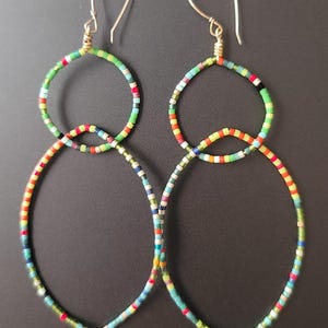 Puede incluir: Un par de pendientes hechos a mano con un diseño único. Los pendientes presentan dos aros conectados, cada uno adornado con cuentas de colores en una variedad de colores, incluyendo verde, rojo, amarillo y azul. Los ganchos para las orejas son dorados.