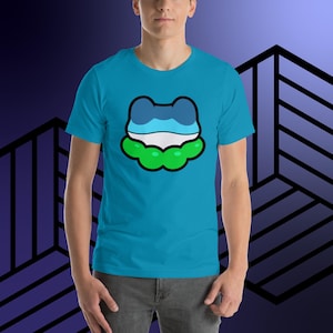 Pode incluir: Uma camiseta turquesa com um desenho de sapo de desenho animado. O sapo tem uma cabeça azul e branca e uma base verde em forma de nuvem, tudo delineado em preto. A camiseta de manga curta é feita de um material macio.