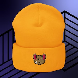 May include: Mustard yellow beanie hat with a folded brim. The hat features an embroidered cartoon mouse face with pink ears and a bandage. The hat is made of a soft, knitted material.