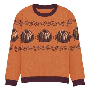 May include: An orange sweater with a crew neck and long sleeves. The sweater features a repeating pattern of brown pumpkins and decorative swirls. The cuffs and neckline are a dark maroon color.