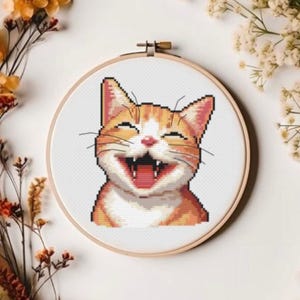 Op de afbeelding: Een kruissteek-borduurring met een close-up van een glimlachende oranje tabby-kat met witte markeringen. De kat heeft zijn mond open en laat zijn tanden zien.