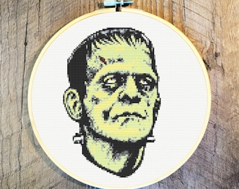 Puede incluir: Una bordado de punto de cruz de la cara del monstruo de Frankenstein. El monstruo tiene piel verde, cabello negro y una cicatriz en la frente. El bordado está enmarcado en un aro de madera redondo.
