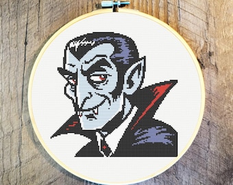 May include: A cross-stitch embroidery hoop displaying a cartoon vampire with red eyes, a black suit, and a red-lined cape. The vampire has a sinister grin and pointed ears. The embroidery is set against a white background.