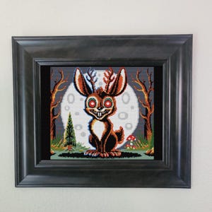 May include: A cross-stitch embroidery of a cartoon rabbit with antlers, red eyes, and a toothy grin. The rabbit is standing in a forest setting with a full moon in the background.
