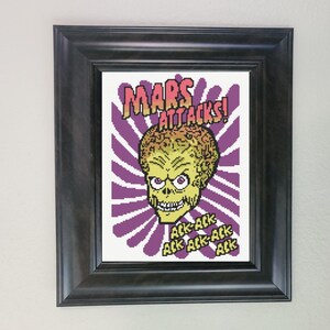 May include: Framed cross-stitch art depicting a green alien with a large head and red eyes, set against a purple and white burst design. The text "MARS ATTACKS!" is above the alien, with "Ack Ack Ack" below. The frame is black.