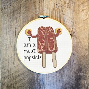 May include: Cross-stitch art in a round hoop. The design features a meat popsicle with arms and a stick, and the text "I am a meat popsicle". The popsicle is brown and beige, with yellow circles as hands. The background is white.