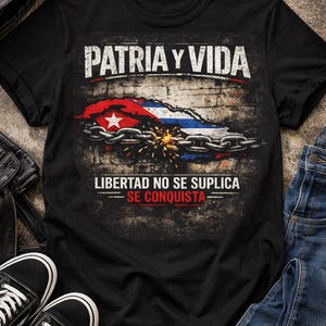 Puede incluir: Camiseta negra con el texto "PATRIA Y VIDA" sobre un gráfico de la bandera cubana, parcialmente encadenada y chispeante. Debajo, el texto dice "LIBERTAD NO SE SUPLICA - SE CONQUISTA".