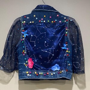 Könnte beinhalten: Jeansjacke mit dunkelblauem Samt-Rückenteil mit Sternbild-Design. Die Jacke ist mit bunten sternförmigen Verzierungen, transparenten Ärmeln und floralen Akzenten verziert. Die Jacke hängt an einem silbernen Kleiderbügel.