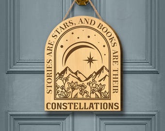 May include: Wooden sign with the text "STORIES ARE STARS, AND BOOKS ARE THEIR CONSTELLATIONS." The sign features a mountain range, flowers, a crescent moon, and a star design. It hangs from a door with a gold door knocker.