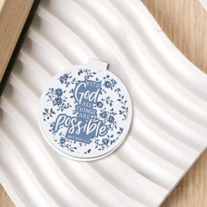 Peut inclure: Un autocollant blanc en forme d'ornement avec un motif floral bleu. L'autocollant porte l'inscription "With God all things are possible" et le verset biblique Matthieu 19:26. Il est posé sur une surface blanche et ondulée.