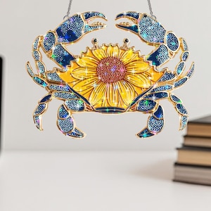 Könnte beinhalten: Dekorative Krabbenverzierung mit Sonnenblumenmitte. Die Krallen und Beine der Krabbe sind blau mit einem gepunkteten Muster, und die Sonnenblume ist gelb mit einem roten Zentrum. Der Schmuck hängt an einer silbernen Kette.