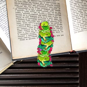 Puede incluir: Un marcapáginas caprichoso con una pila de ranas verdes con detalles de flores rosas. El marcapáginas de rana está colocado sobre un libro abierto, añadiendo un toque de naturaleza y encanto a la lectura.