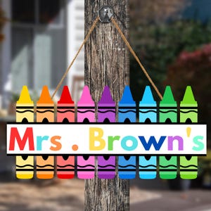 May include: A colorful wooden sign featuring a row of crayon illustrations in various colors. The sign has the text "Mrs. Brown's" in a rainbow font. The sign is suspended by a rope.