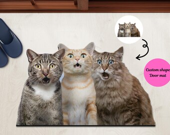 May include: Three cats with surprised expressions on their faces are featured on a custom shape door mat. The mat is black and the cats are in a row, with the cat on the left being a tabby, the middle cat being a ginger, and the cat on the right being a long-haired cat.