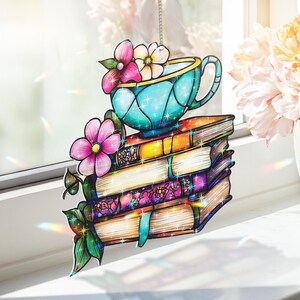 Puede incluir: Colgante decorativo para ventana con una pila de libros, una taza de té turquesa y flores. Los libros tienen cubiertas naranjas, moradas y con motivos florales. Flores rosas y blancas y hojas verdes adornan el diseño, que cuelga de una cadena.