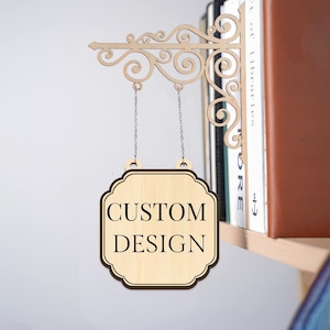 Pode incluir: Uma placa de madeira com as palavras "CUSTOM DESIGN" em letras pretas. A placa é suspensa por correntes de um suporte decorativo com volutas. O suporte está fixado a uma prateleira com livros.