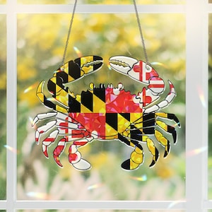 May include: A clear acrylic crab-shaped ornament featuring the Maryland state flag design. The flag is a black and gold chequered pattern with a red and white cross in the top left corner.