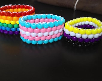 May include: Three colorful beaded bracelets. One features a rainbow pattern, another has pink, white, and light blue beads, and the third has yellow, white, purple, and black beads. The bracelets are stacked and cylindrical.