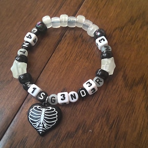 May include: A beaded bracelet with a black heart-shaped charm featuring a white skeleton design. The bracelet has black, white, and clear beads, with some beads displaying letters and numbers. The bracelet is on a wooden surface.