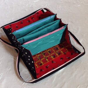 May include: A black zippered pouch with a pink zipper and a red, black, and gold patterned fabric lining. The pouch has four separate compartments with zippers.