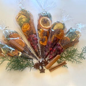 Peut inclure: Plusieurs cônes en cellophane transparent remplis de tranches d'orange séchées, de bâtons de cannelle, d'anis étoilé et de baies rouges. Des brins verts et de l'anis étoilé sont disposés autour de la base des cônes. Les cônes sont attachés en haut avec des attaches torsadées transparentes.