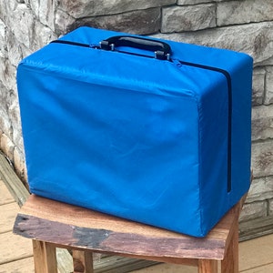 Puede incluir: Una funda de transporte azul con cremallera y asa negra. La funda está sobre un taburete de madera.