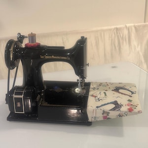 Może przedstawiać: Czarna maszyna do szycia Singer w stylu vintage ze złotym napisem The Singer Manufacturing Co. Maszyna posiada silnik, szpulkę nici na g&oacute;rze oraz dołączony st&oacute;ł przedłużający pokryty tkaniną z nadrukiem o tematyce krawieckiej.