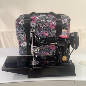 Pode incluir: Uma máquina de costura Singer preta vintage com detalhes dourados e um carretel de linha prateado no topo. Atrás da máquina, há uma mala de transporte retangular de tecido acolchoado com um padrão floral escuro de flores rosas e brancas.