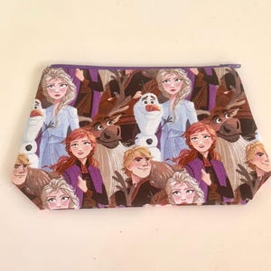 May include: A rectangular cosmetic bag featuring a repeating pattern of characters from the animated film "Frozen." The bag has a purple zipper and a light-colored background. The characters include Elsa, Anna, Olaf, Kristoff, and Sven.