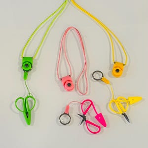 Puede incluir: Cuatro cordones de colores con diferentes accesorios, incluyendo un par de tijeras, una lupa y un pequeño clip. Los cordones son verdes, amarillos, rosas y amarillos.