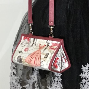 Könnte beinhalten: Eine kleine, rot-weiß gemusterte Handtasche mit einem burgunderfarbenen Riemen. Die Tasche hat einen Klappverschluss und ein Design mit einer Frau in einem Kleid und Nähwerkzeugen.