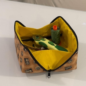 May include: A brown fabric zippered pouch with a yellow lining. The pouch is open and contains a green spool of thread, a rotary cutter, and a green and yellow seam ripper.