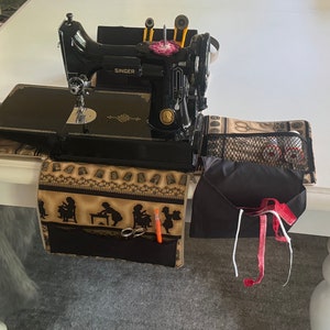 Pode incluir: Uma máquina de costura Singer preta com uma mesa dobrável e uma bolsa de armazenamento de tecido preta com um bolso de malha. A máquina de costura está sentada em uma mesa branca com um corredor de tecido marrom e preto com um padrão de meninas costurando.