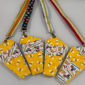 Puede incluir: Cinco porta tijeras de tela amarilla con patrones de abejas y costura. Cada porta tijeras tiene una correa de tela con un color y un patrón diferente.