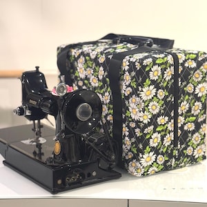 Puede incluir: Una máquina de coser Singer negra con una funda de transporte de estampado de margaritas blancas y negras. La funda tiene una cremallera y un asa.