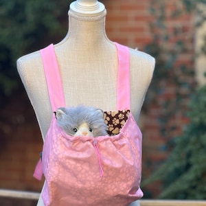 May include: A pink pet carrier backpack with paw print design. A gray plush toy cat is inside the carrier.