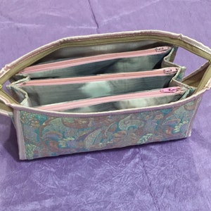 May include: A multi-compartment zippered pouch with a paisley pattern in shades of pink, green, and gold. The pouch has three separate zippered compartments.