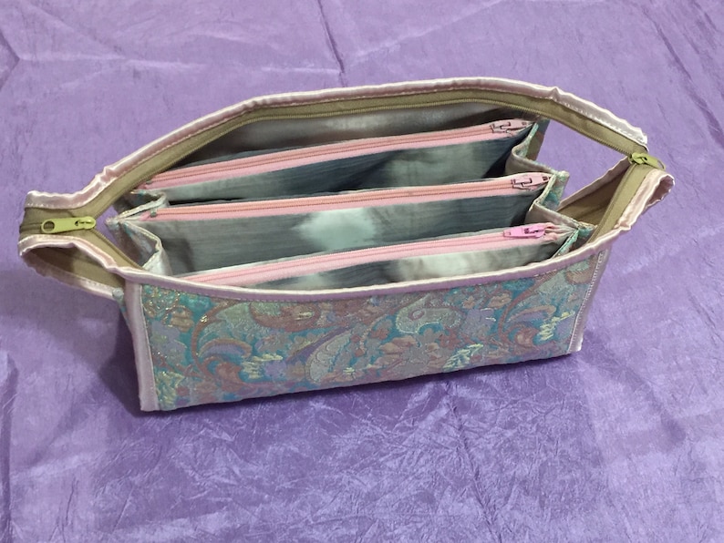 May include: A multi-compartment zippered pouch with a paisley pattern in shades of pink, green, and gold. The pouch has three separate zippered compartments.