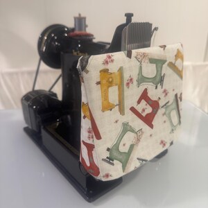 Może przedstawiać: Czarna maszyna do szycia częściowo przykryta prostokątnym pokrowcem z tkaniny w powtarzający się wz&oacute;r maszyn do szycia w stylu vintage w kolorach ż&oacute;łtym, czerwonym i zielonym, z akcentami małych r&oacute;żowych kwiat&oacute;w na kremowym tle w kratkę.