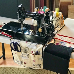 Peut inclure: Une machine à coudre Singer noire avec une housse de table de machine à coudre en tissu blanc et marron avec des poches et une pochette en tissu noir. La housse de table a un motif de machines à coudre et le mot "Singer" imprimé dessus.