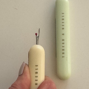 May include: Two small, handheld threaders with red tips. The threader on the left is cream colored with the text "THREAD A NEEDLE" printed on the side. The threader on the right is light green with the text "THREAD A NEEDLE" printed on the side.