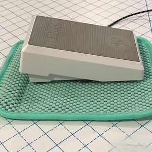 May include: A white sewing machine foot pedal with a gray top and the word "BERNINA" printed on it. The pedal is sitting on a light green non-slip mat.